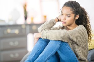 a lady with poor mental health.  A teenage girl of African descent is indoors in a bedroom. She is wearing casual clothing. She is sitting on her bed and looking sad.